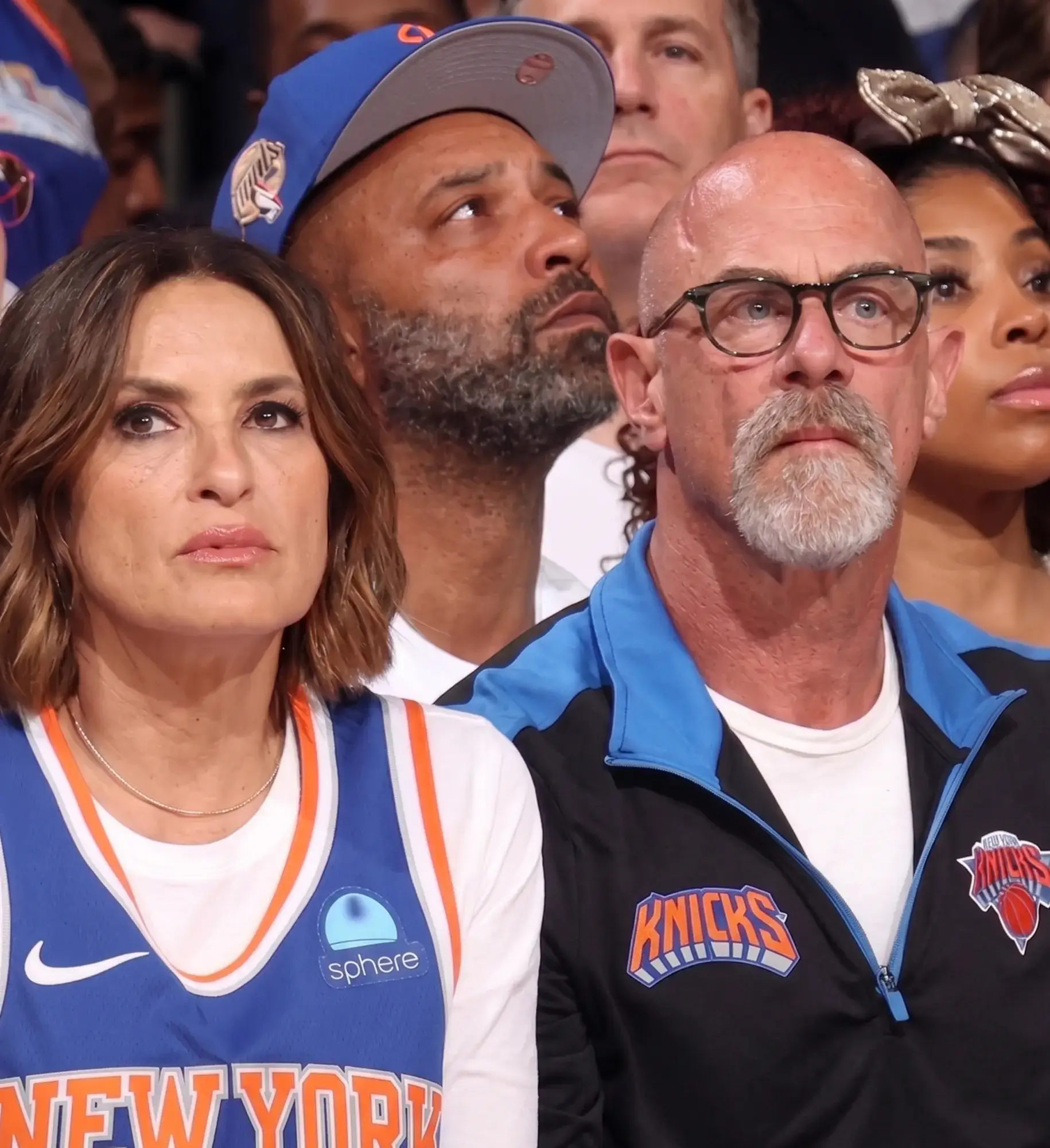 Mariska Hargitay in a blue New York Knicks jersey sits next to Christopher Meloni wearing a black and blue Knicks zip-up jacket at a basketball game.