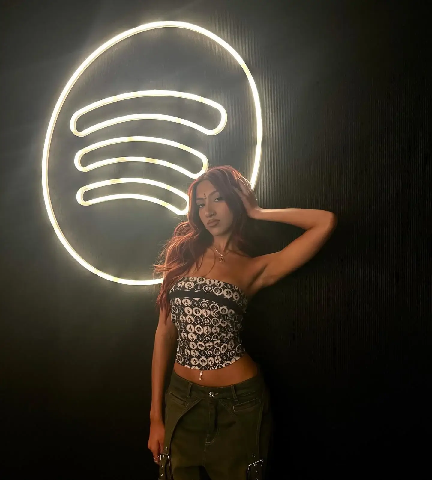 Mariska Hargitay with reddish-brown hair poses in a patterned strapless tube top and cargo pants in front of a glowing neon Spotify logo.