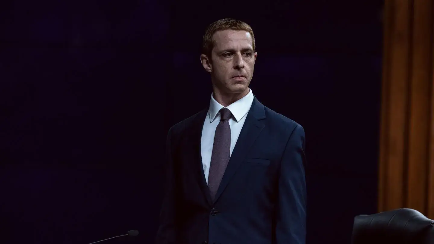 Jeremy Strong stands in a dark suit, white button-down shirt, and brown tie while looking off-camera against a dark background.