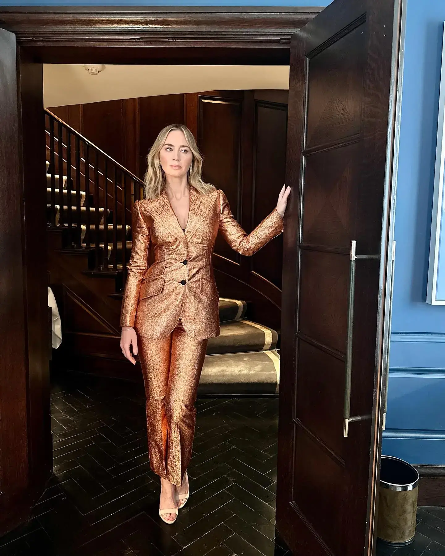 Emily Blunt stands in a doorway wearing a shimmering copper pantsuit and strappy heels with a wooden staircase visible behind her.