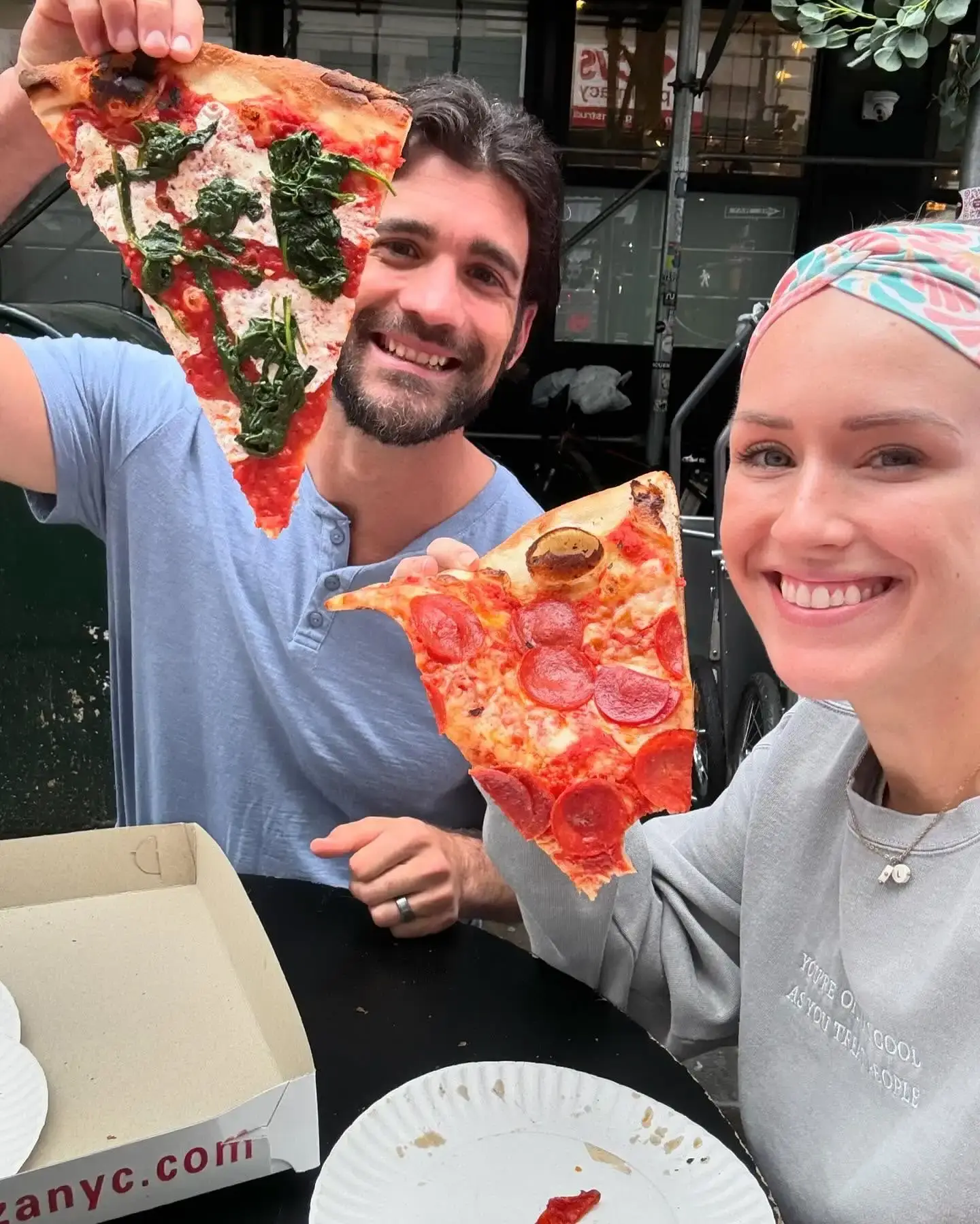 Jen Hamilton in a colorful headband and gray sweatshirt smiles next to her ex-husband Brian Hamilton as they both hold up large slices of pizza.