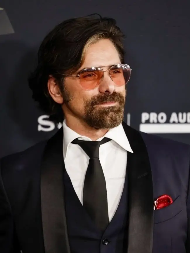 John Stamos poses with a full beard, mustache, and tinted aviator glasses while wearing a sleek tuxedo with a red pocket square.