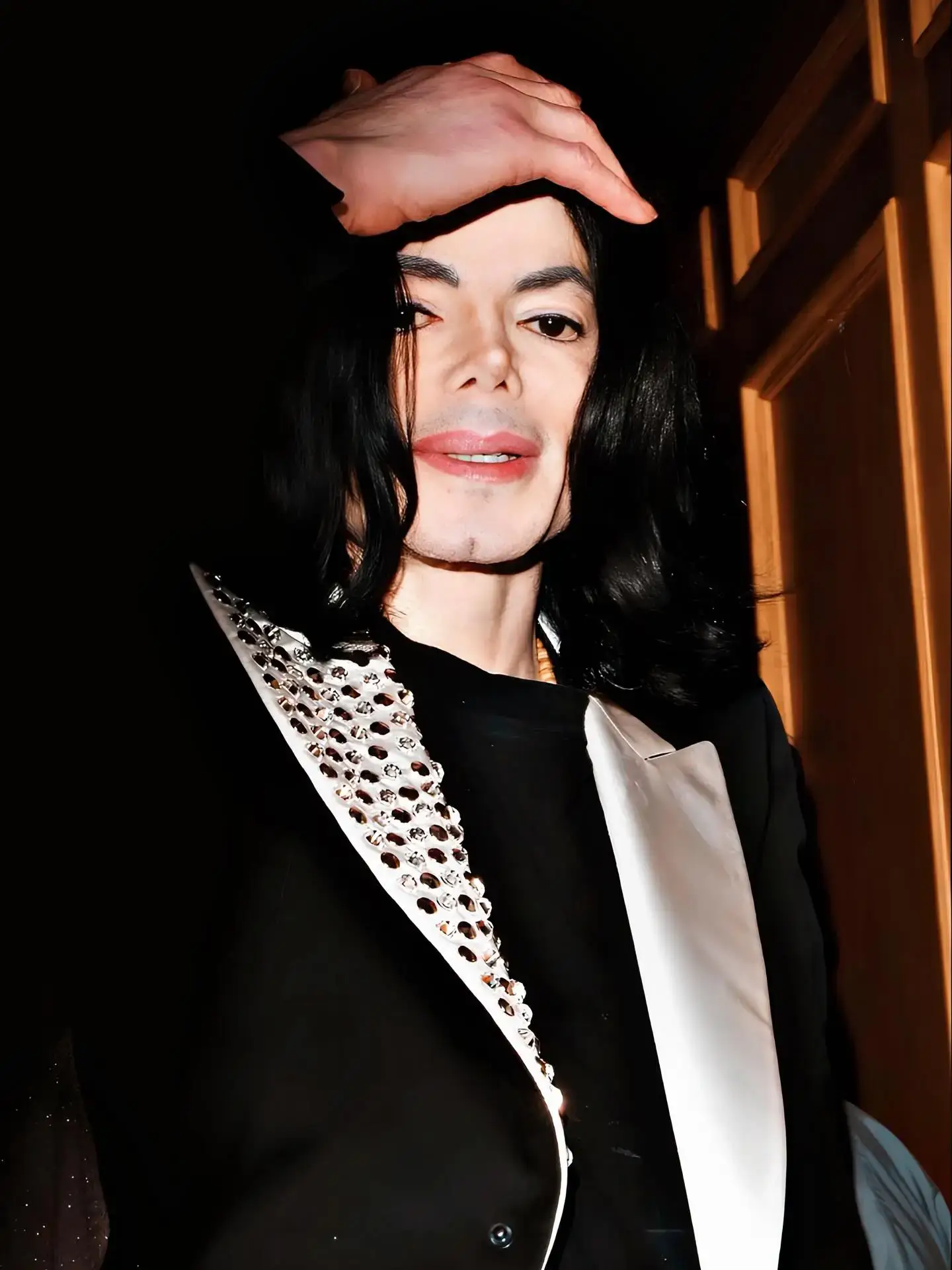 Michael Jackson holds his hand near his forehead while smiling, wearing a black jacket with heavily studded white lapels.