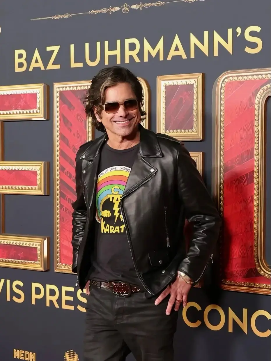 John Stamos smiles in a black leather jacket, sunglasses, and a colorful graphic t-shirt in front of a "BAZ LUHRMANN