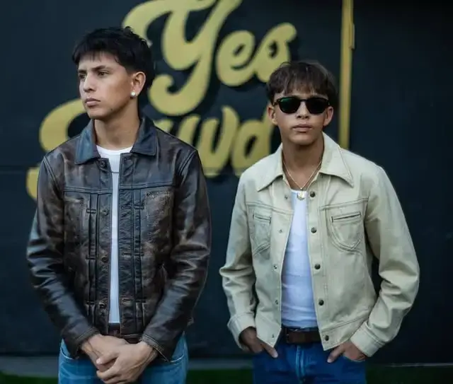 Nyjah Music in a distressed brown leather jacket stands next to Zyah Rhythm wearing sunglasses and a cream jacket in front of a blurred "Yes" logo.
