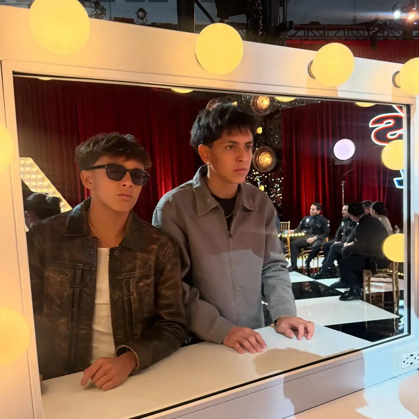 Zyah Rhythm in a leather jacket and sunglasses stands with Nyjah Music in a gray jacket, captured as a reflection in a lit vanity mirror with a stage set visible behind them.