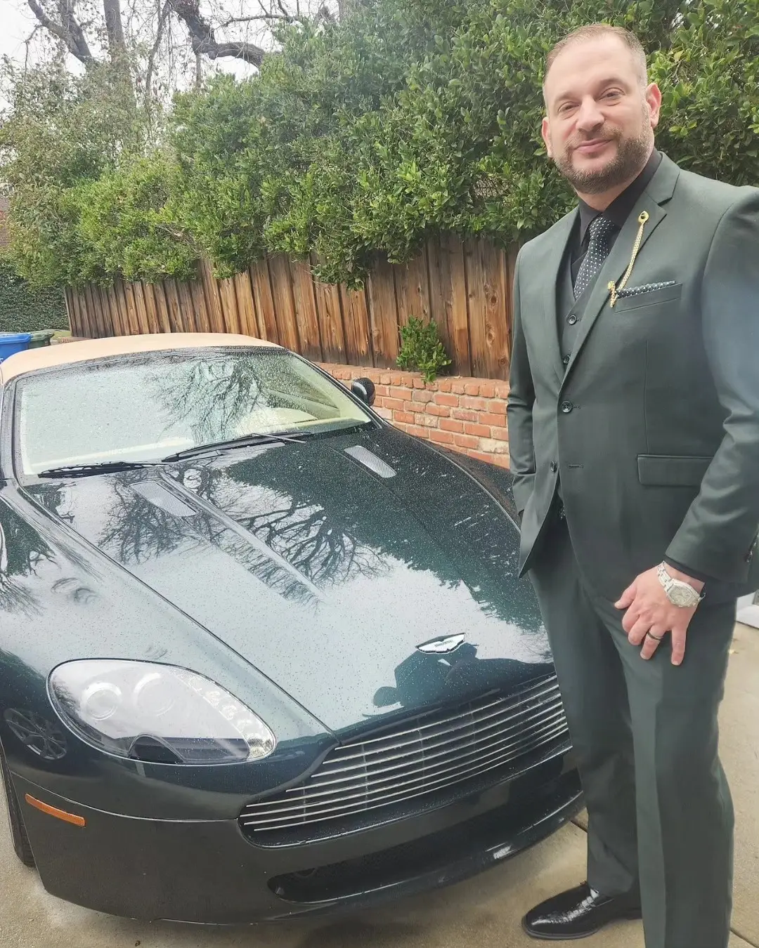 Matthew “M80” Markoff stands in a sharp dark green three-piece suit next to a sleek, dark green Aston Martin luxury sports car.