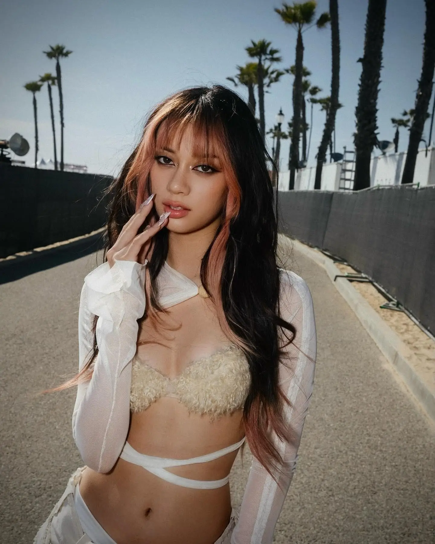 Megan Skiendiel poses outdoors on a sunlit path lined with palm trees, wearing a white mesh top, a textured beige bralette, and strappy details.