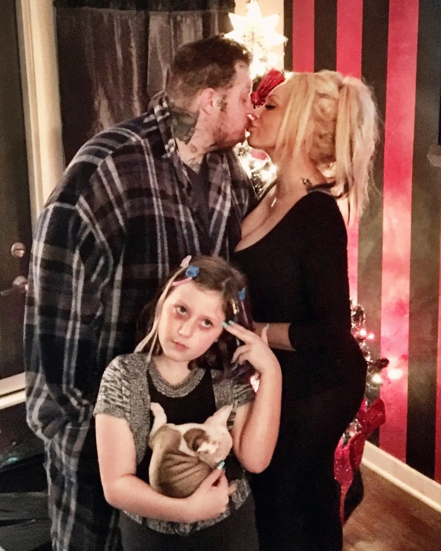 Alt Text: Jelly Roll in a plaid shirt and Bunnie XO in a black dress kiss in front of a Christmas tree while their daughter holding a small dog makes a funny face in the foreground..