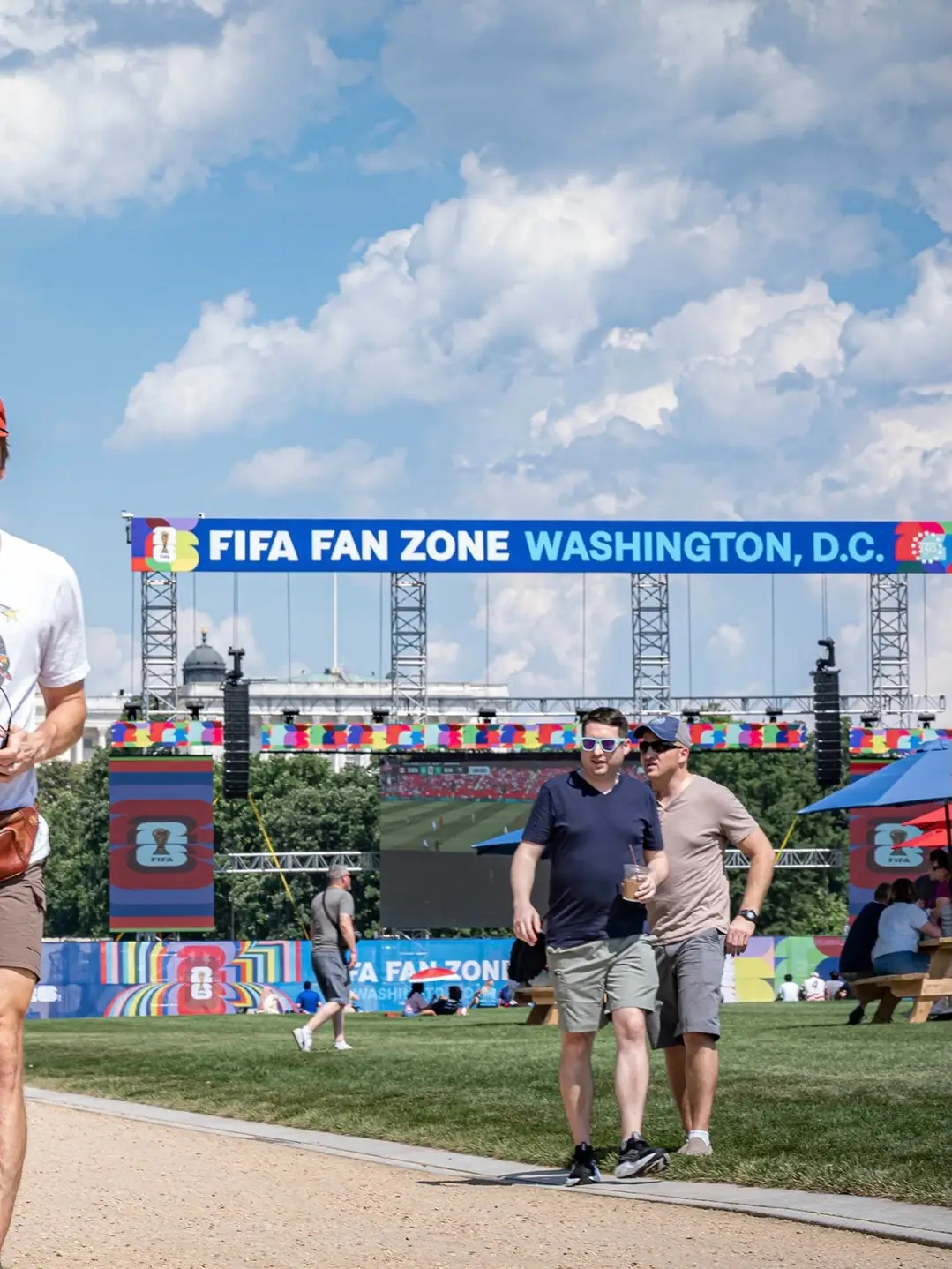 People walk across a sunny lawn in front of a large outdoor stage setup featuring a blue banner that reads "FIFA FAN ZONE WASHINGTON, D.C.".