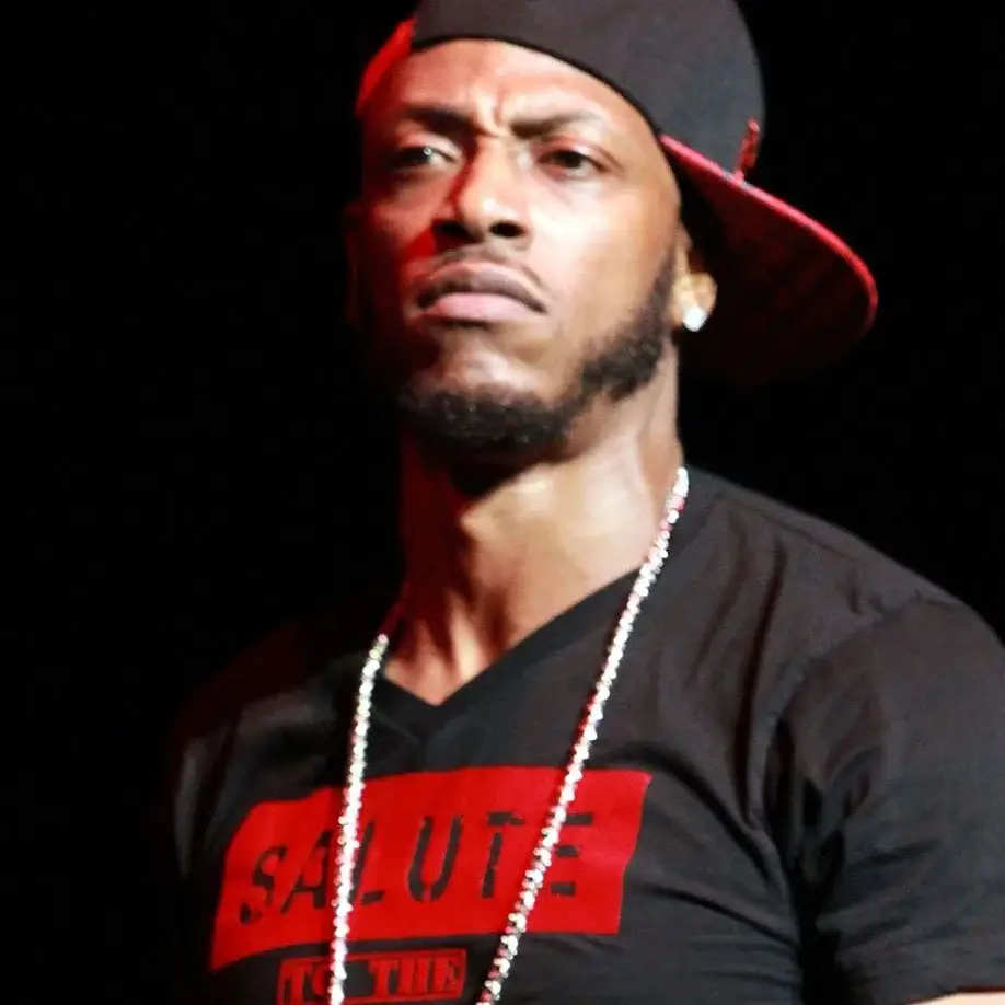 Rapper Mystikal looks to the side with a serious expression, wearing a sideways black and red cap, a diamond chain, and a black t-shirt with a red logo.