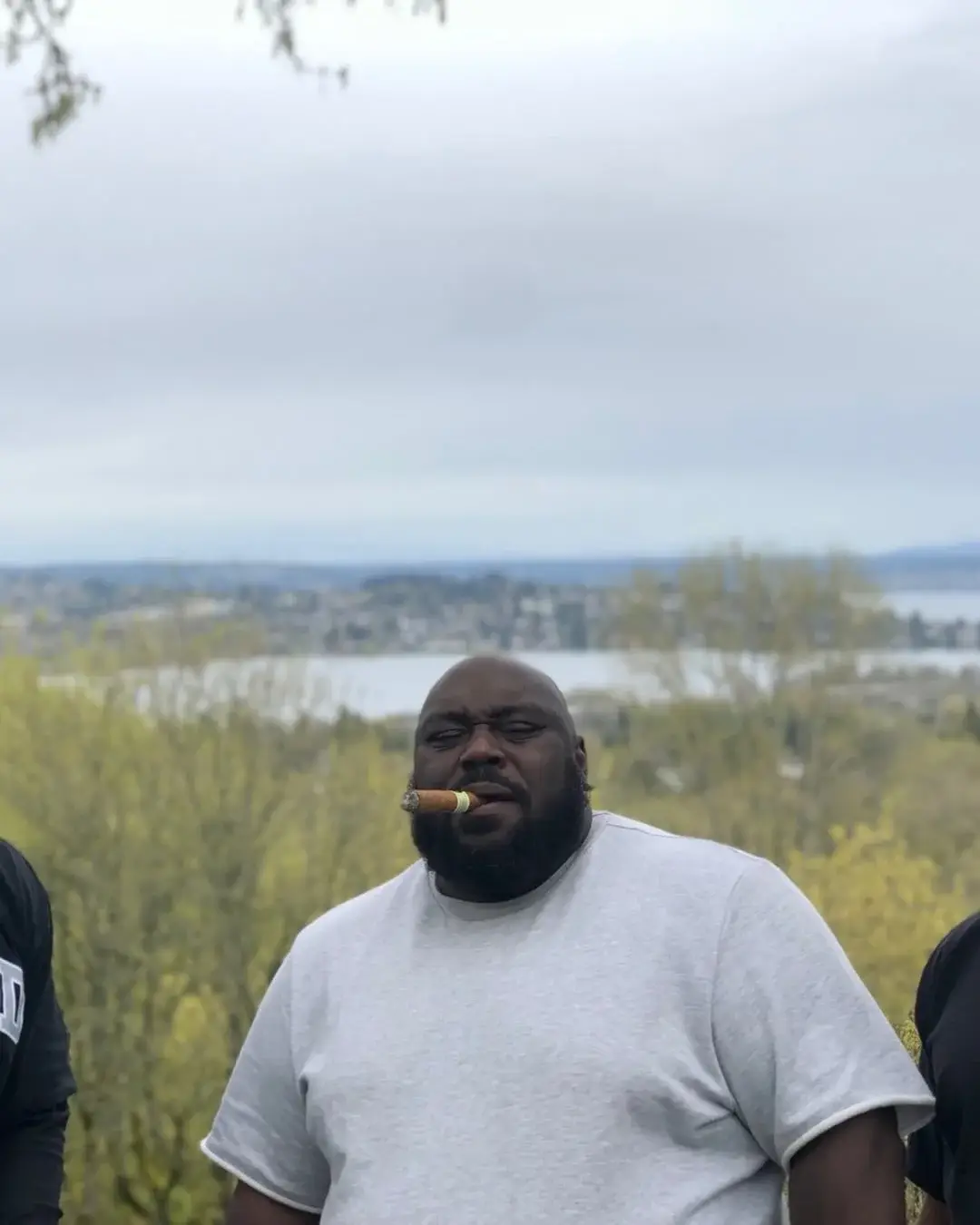 Actor and comedian Faizon Love closes his eyes while enjoying a cigar outdoors, with a scenic backdrop of trees, a lake, and a distant city skyline under an overcast sky.