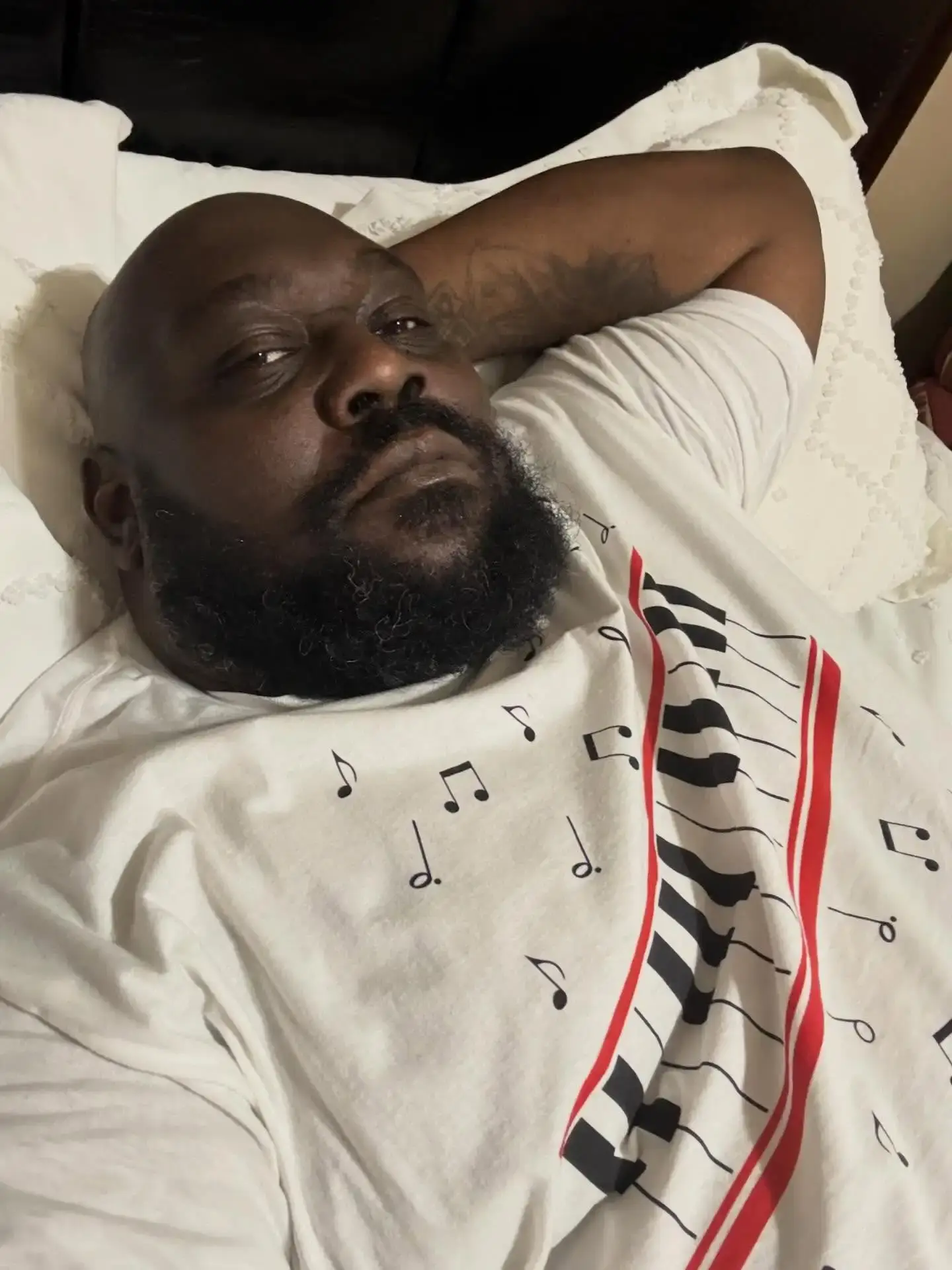 Comedian Faizon Love relaxes in bed with his arm behind his head while wearing a white t-shirt featuring piano keys and musical notes.
