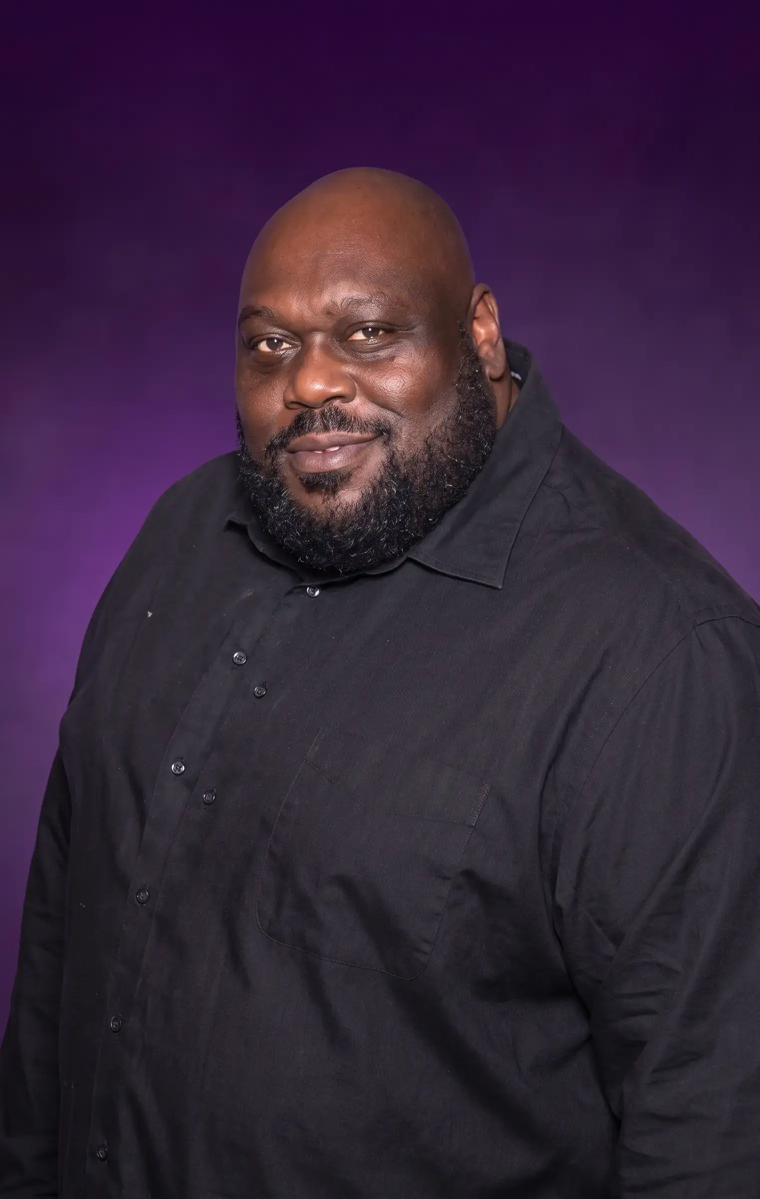 Actor and comedian Faizon Love smiles for a studio portrait wearing a black button-up shirt against a vibrant purple background