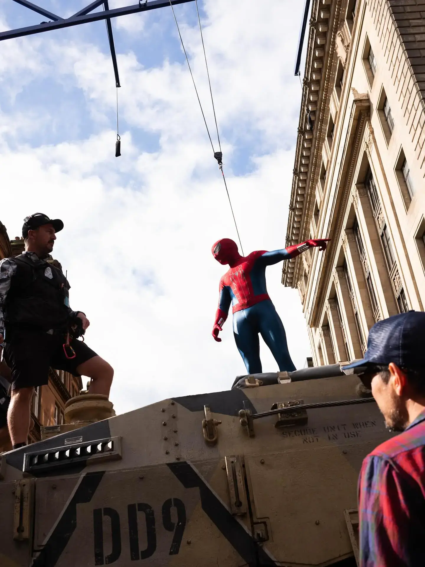An actor in a classic red and blue Spider-Man suit stands harness-attached atop a military tank and points to the distance on the city streets movie set of Spider-Man: Brand New Day.