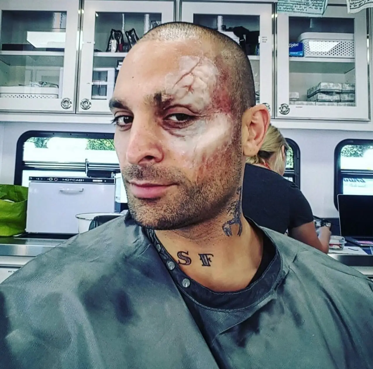 Actor Michael Mando smirks in a makeup chair, sporting intense facial scar prosthetic makeup and a scorpion neck tattoo for his role as Mac Gargan.