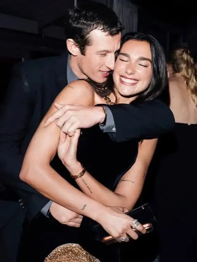 Callum Turner hugs his wife Dua Lipa from behind while she smiles radiantly, holding a small black clutch during a celebratory indoor evening event.