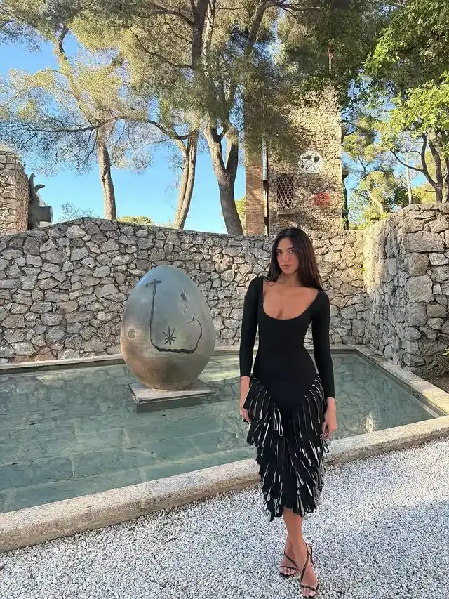 Dua Lipa poses outdoors in a low-cut black long-sleeve dress with unique layered vertical ruffles at the skirt, standing by a stone water fountain pool.