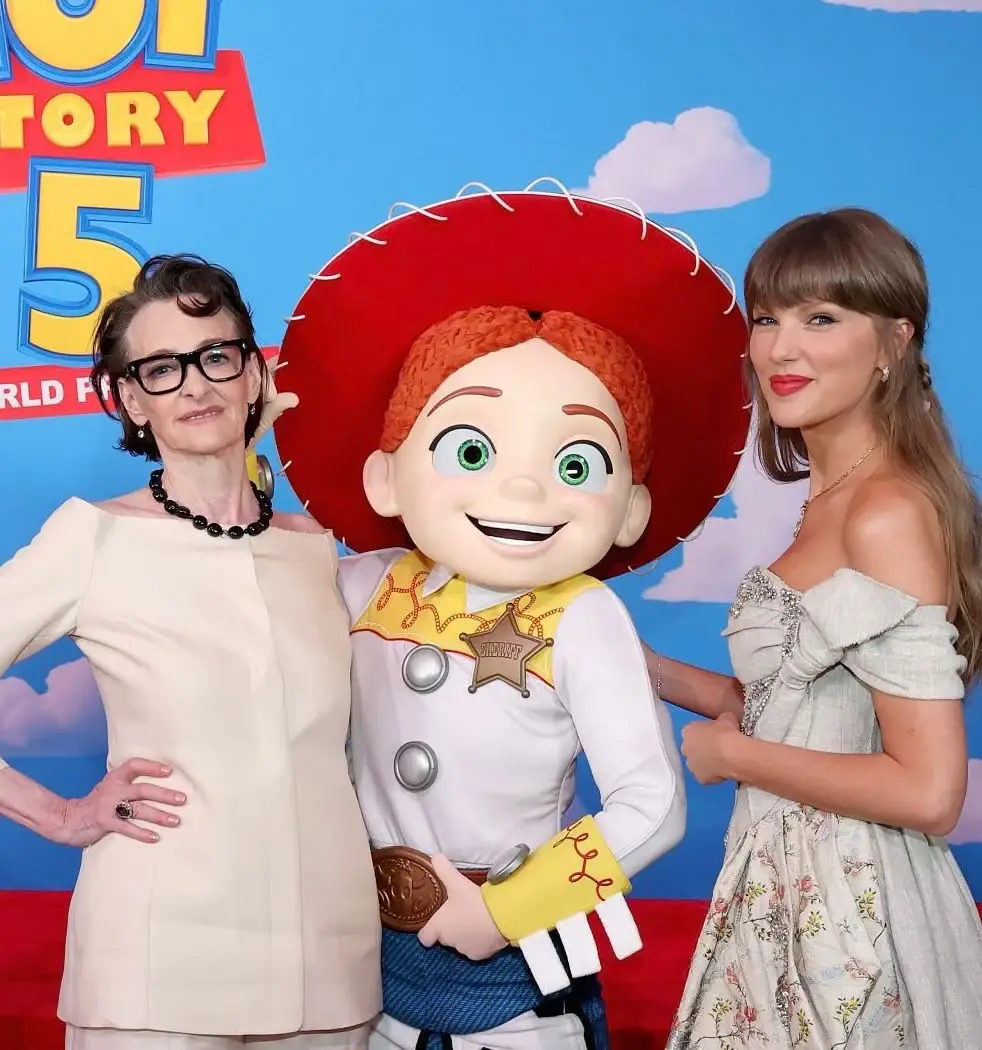 Actress Joan Cusack and singer Taylor Swift pose together on a red carpet on either side of a costumed Jessie character from Toy Story,