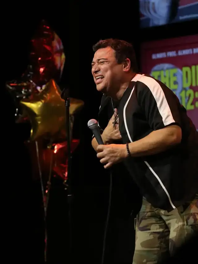 Comedian Carlos Mencia holds a microphone and grimaces expressively with his hand over his chest while performing stand-up comedy on a dark stage.