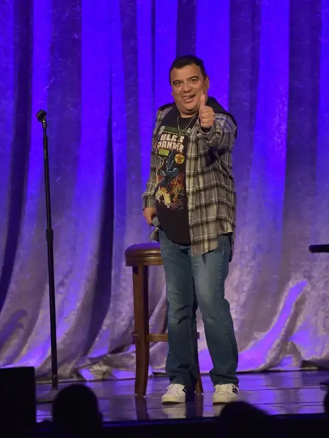 Comedian Carlos Mencia stands on a stage giving a thumbs-up while wearing a plaid shirt and a graphic t-shirt against a purple curtain backdrop.