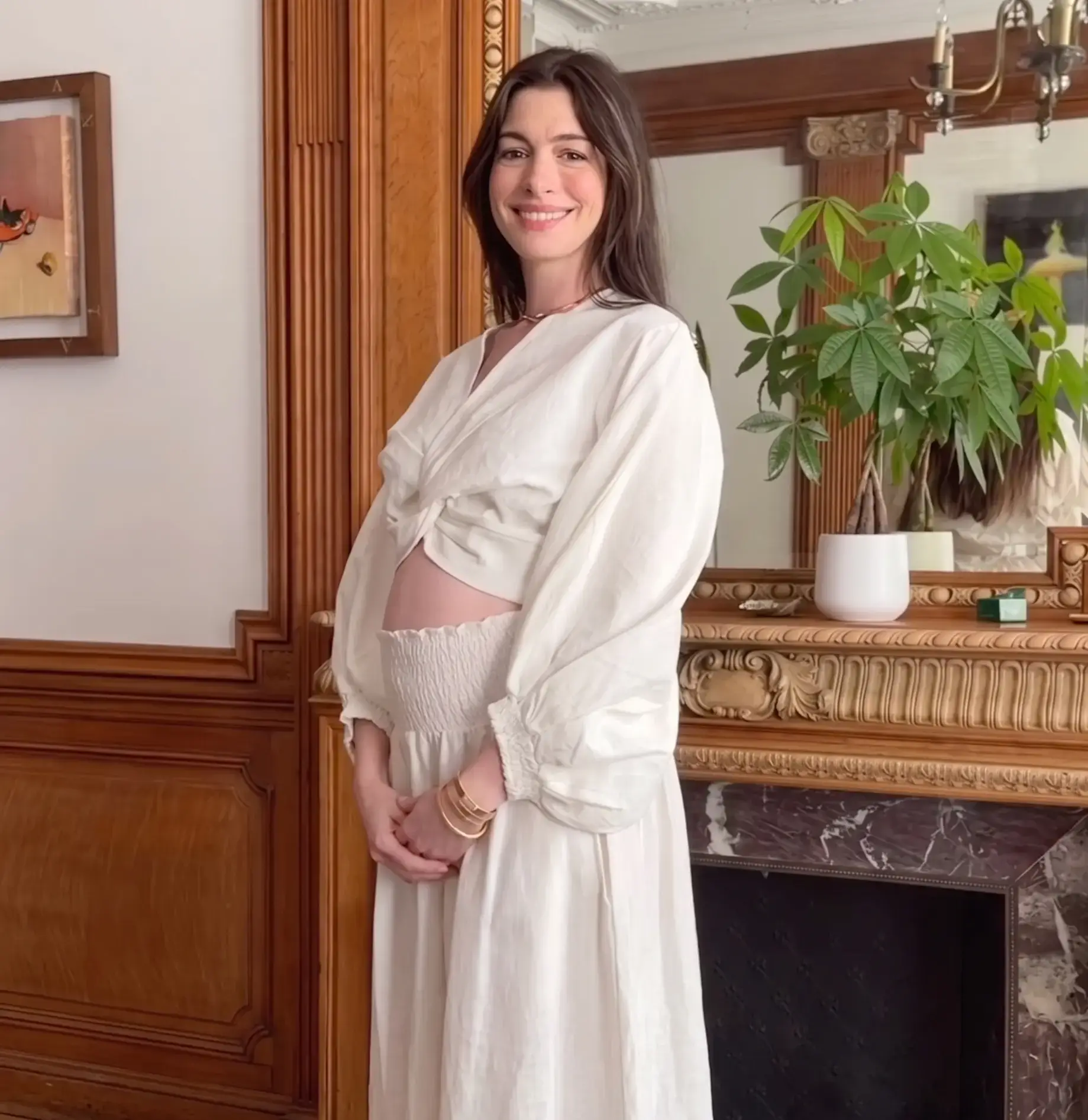Actress Anne Hathaway smiles warmly in a matching white linen crop top and maxi skirt, showing off her pregnant baby bump in front of a wood-paneled fireplace