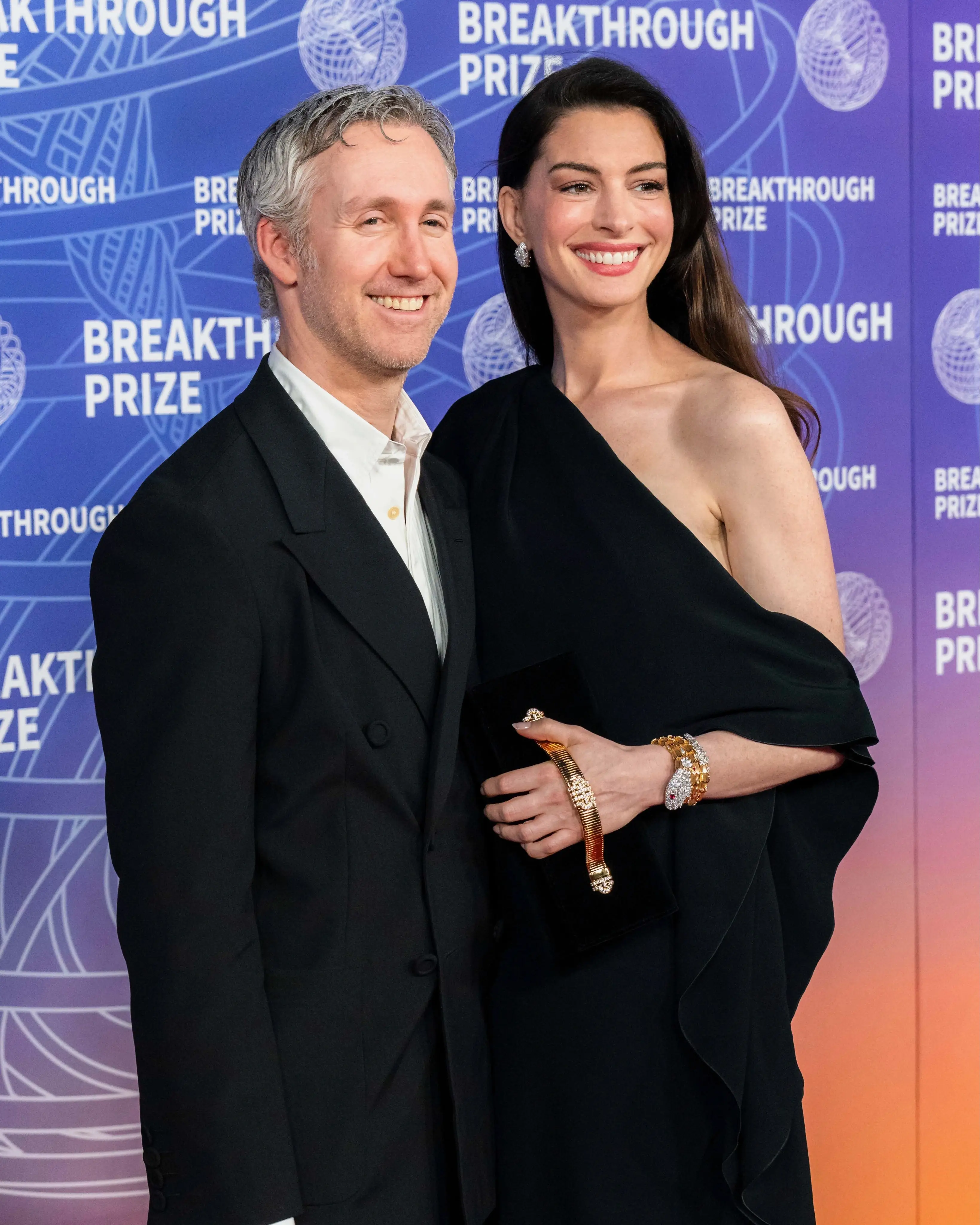 Anne Hathaway, in a glamorous black one-shoulder gown, smiles alongside her husband Adam Shulman on the red carpet at the Breakthrough Prize ceremony.