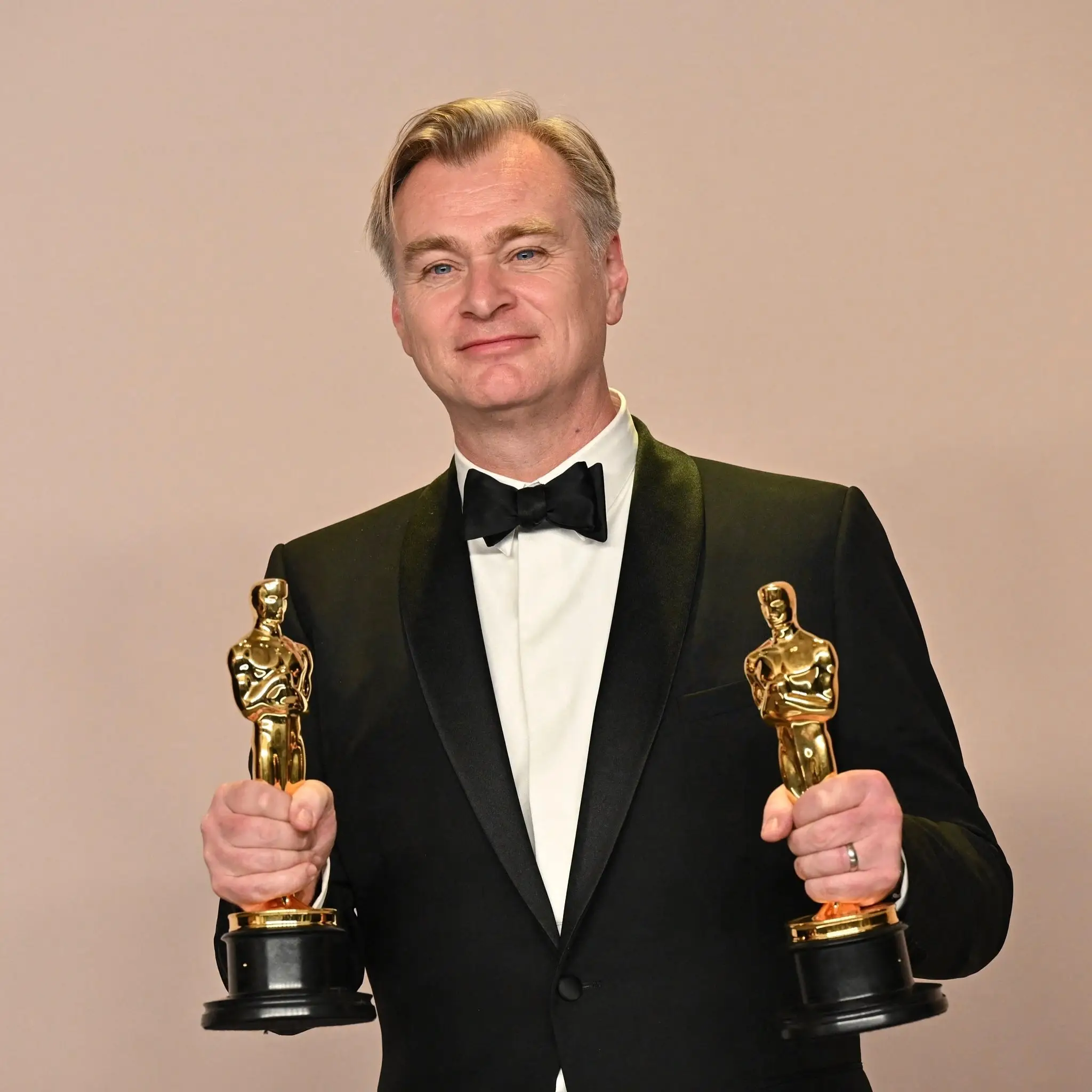 Film director Christopher Nolan smiles proudly while wearing a classic black tuxedo and holding an Academy Award statuette in each hand.