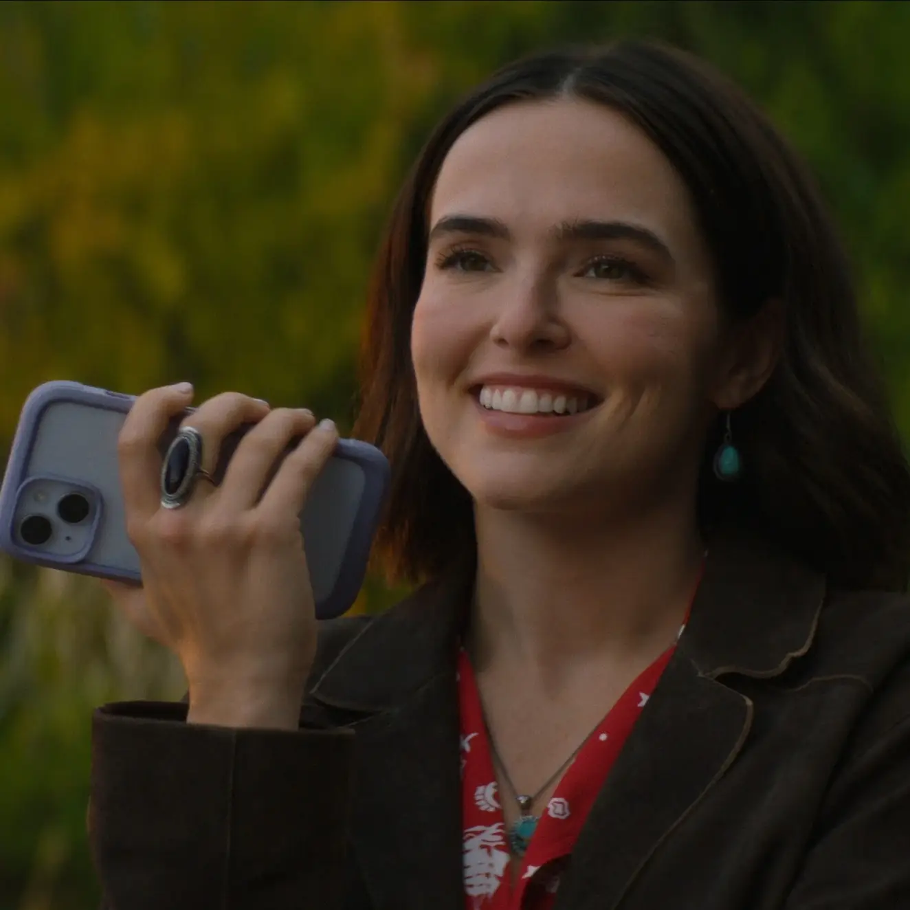 Actress Zoey Deutch smiles brightly while holding a smartphone in a purple case up to her ear outdoors.