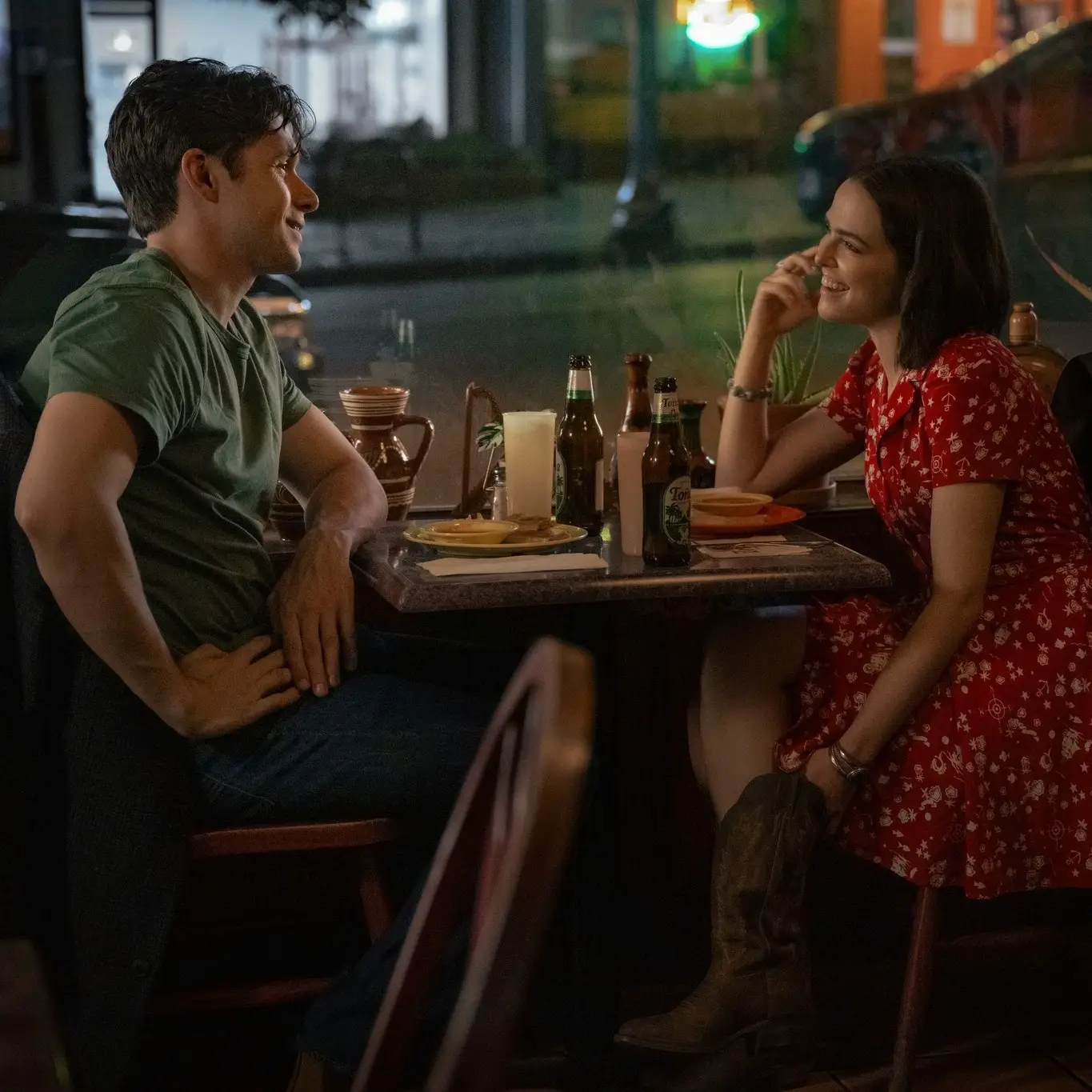 Actors Nick Robinson and Zoey Deutch sit opposite each other at a restaurant table by a window, smiling and talking over a casual meal.