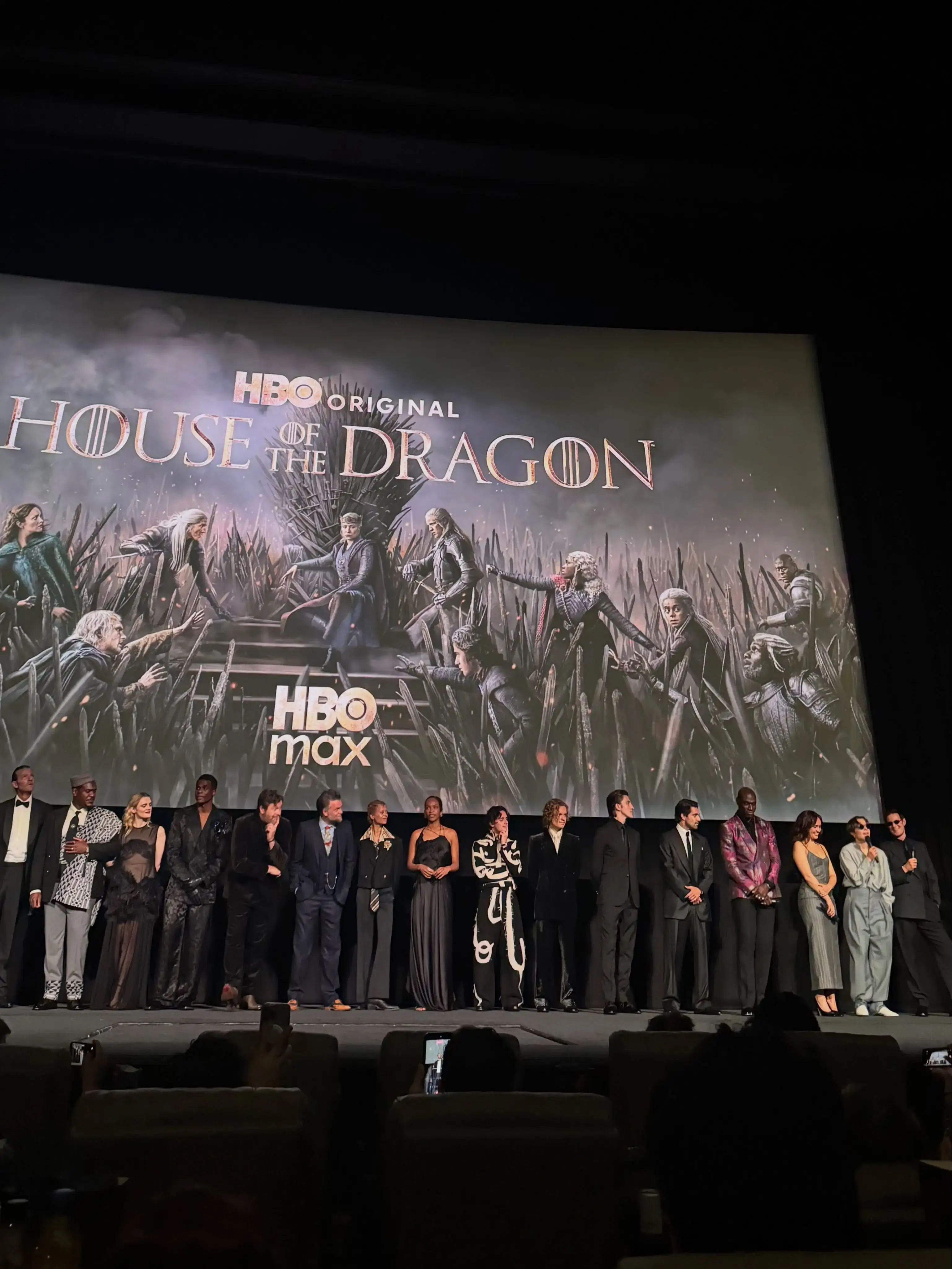 The cast of House of the Dragon stands on a stage in front of a massive "House of the Dragon" and "HBO Max" promotional screen during a premiere event.