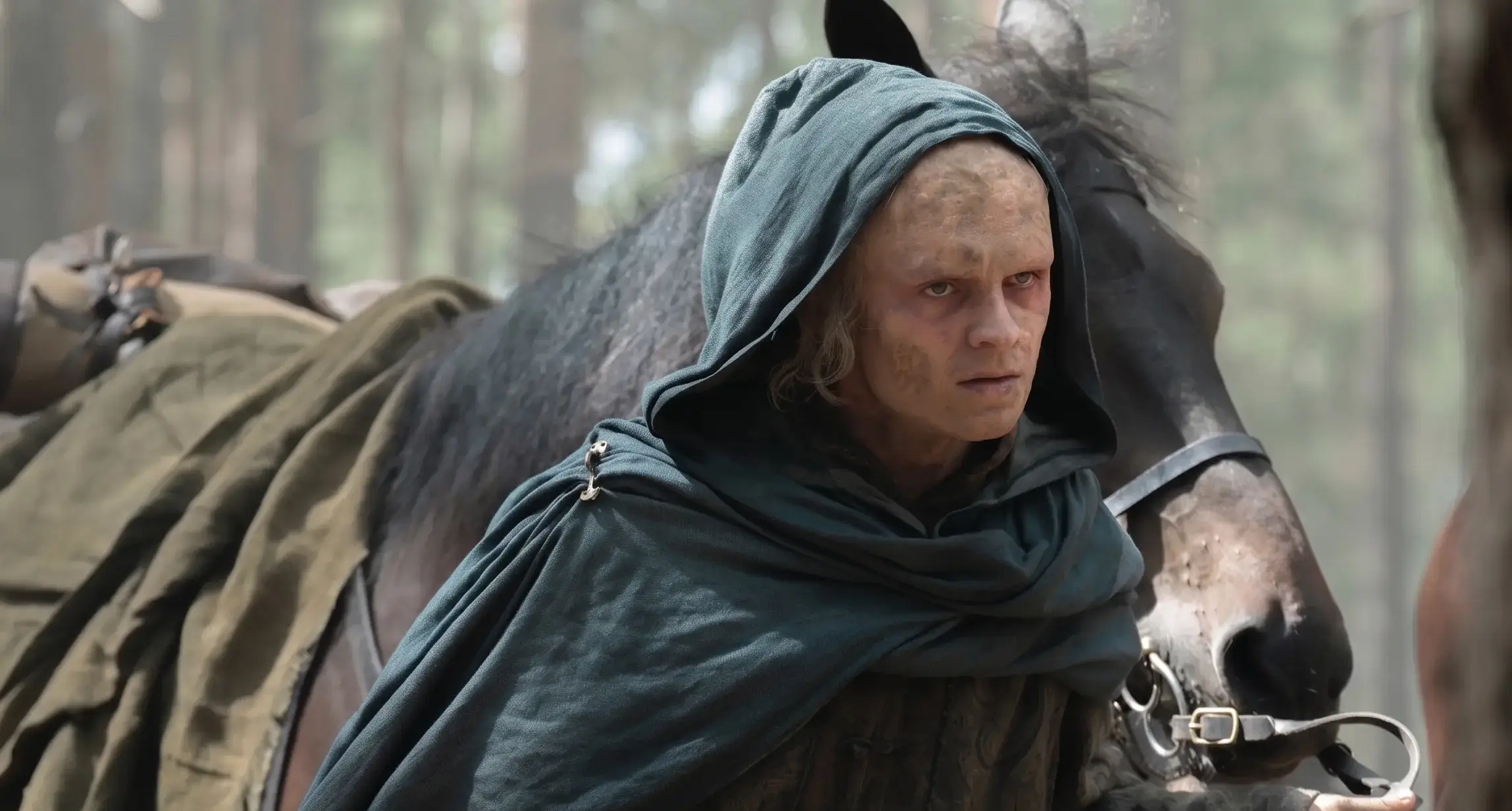 Tom Glynn-Carney, portraying a heavily scarred Aegon II Targaryen in a dark hooded cloak, stands next to a horse in a forest setting.