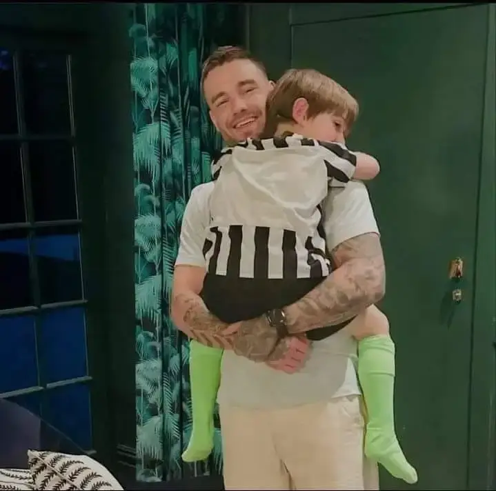 Singer Liam Payne smiles warmly while holding and hugging his young son, Bear, who is wearing a black-and-white striped shirt.
