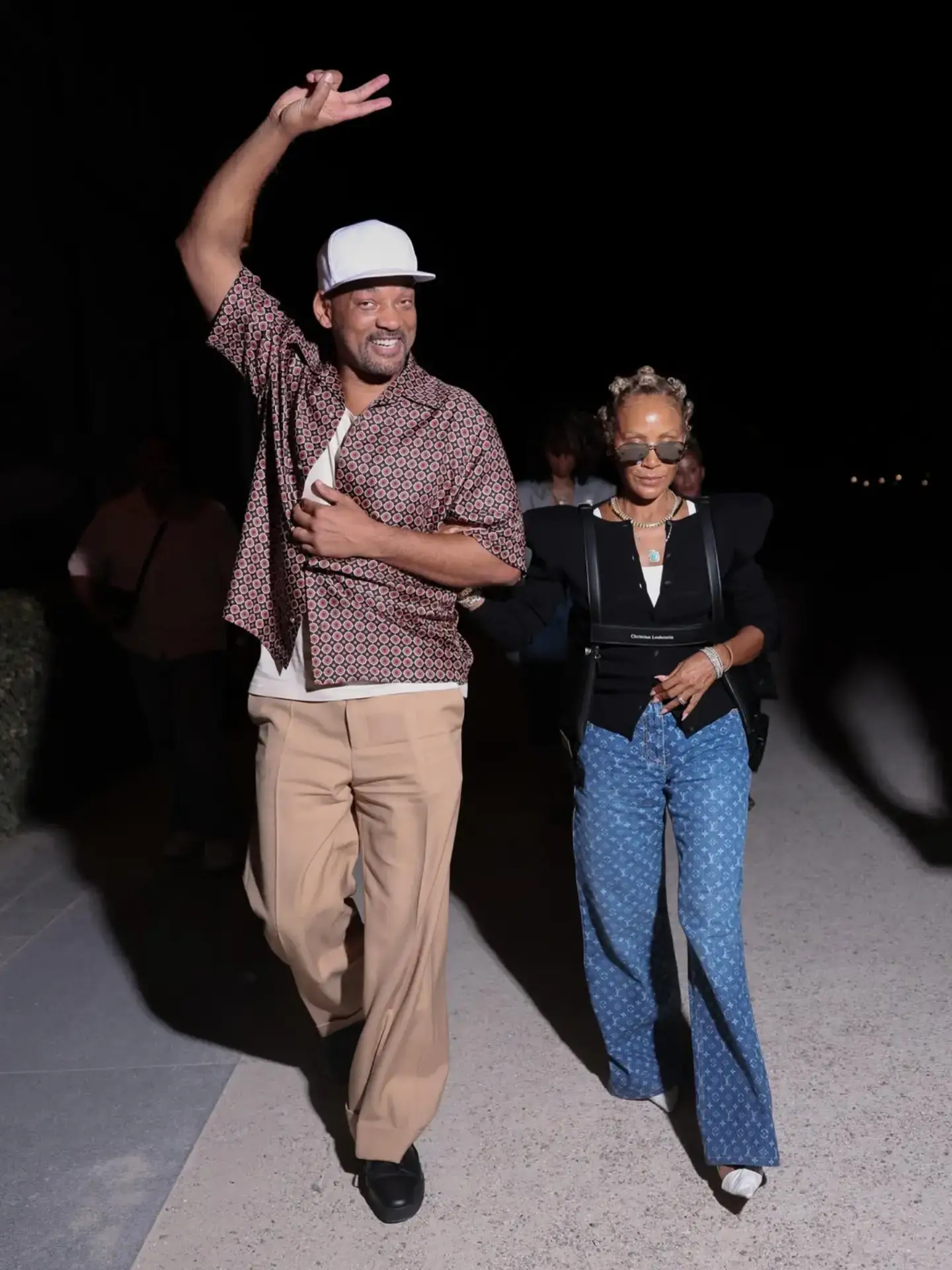 Will Smith waves happily while walking arm-in-arm with Jada Pinkett Smith at night,