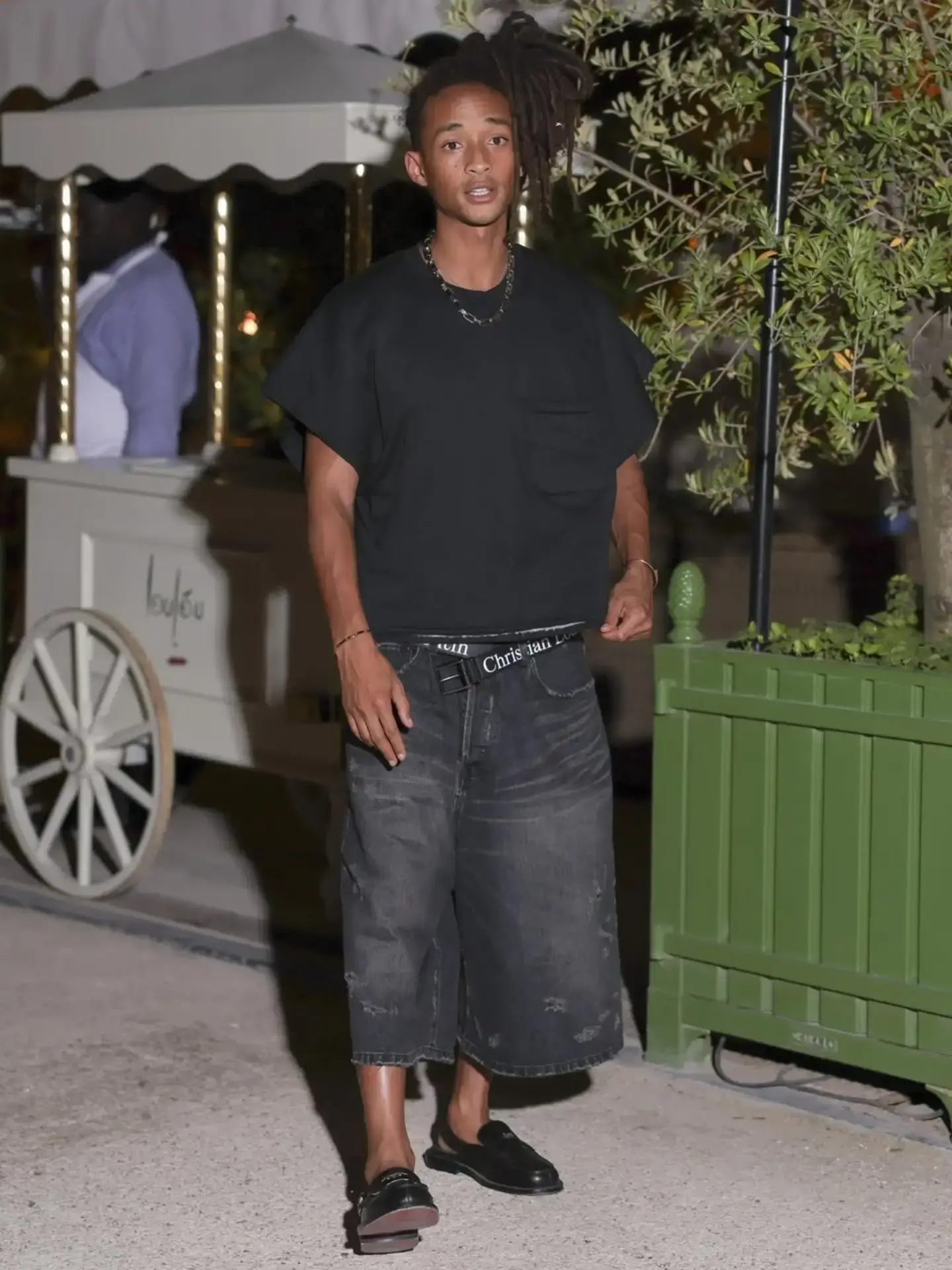 Jaden Smith stands outdoors at night wearing an oversized black t-shirt, baggy denim shorts, and a Christian Louboutin belt.