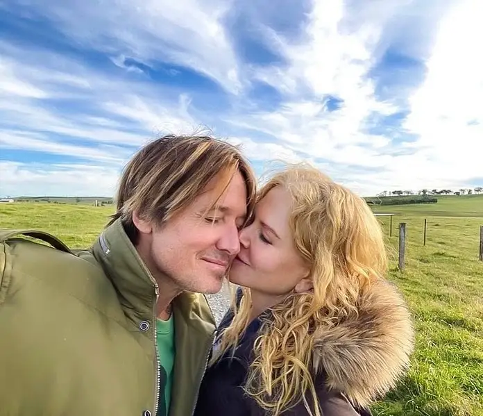 Nicole Kidman kisses Keith Urban on the cheek while standing outdoors in front of a green field under a blue sky.