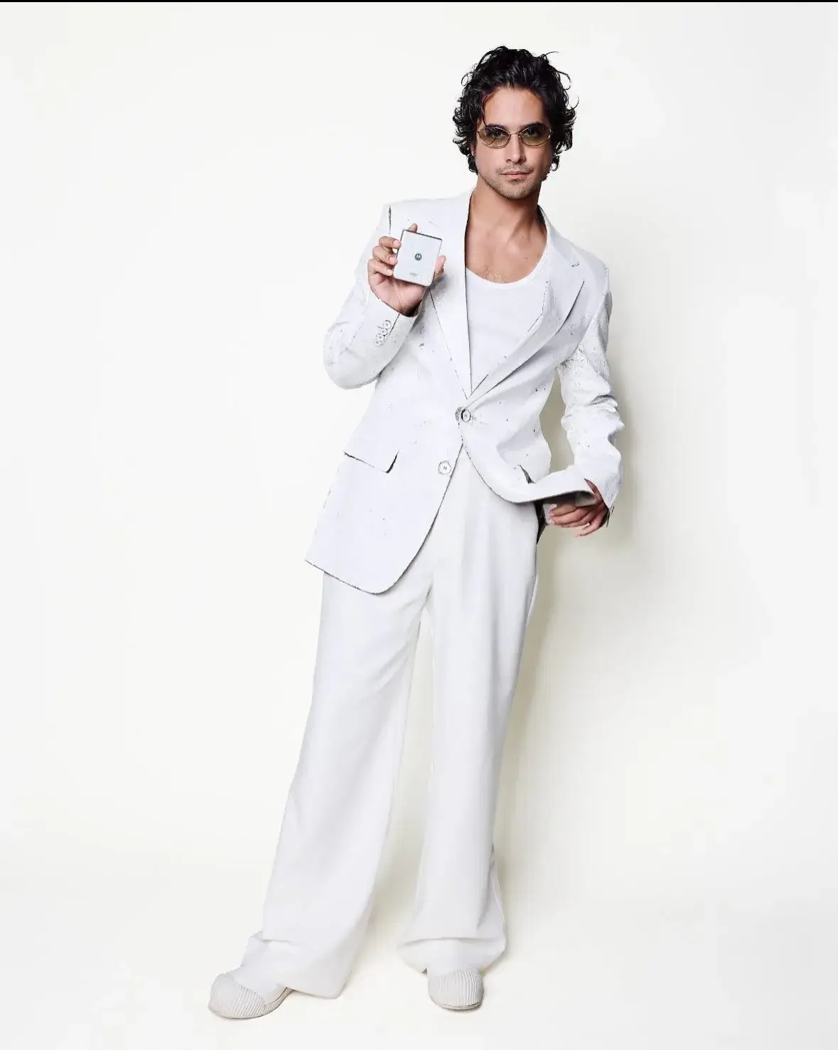 Actor Avan Jogia poses in a stylish all-white suit, tank top, and sunglasses while holding a small electronic device against a plain white background.