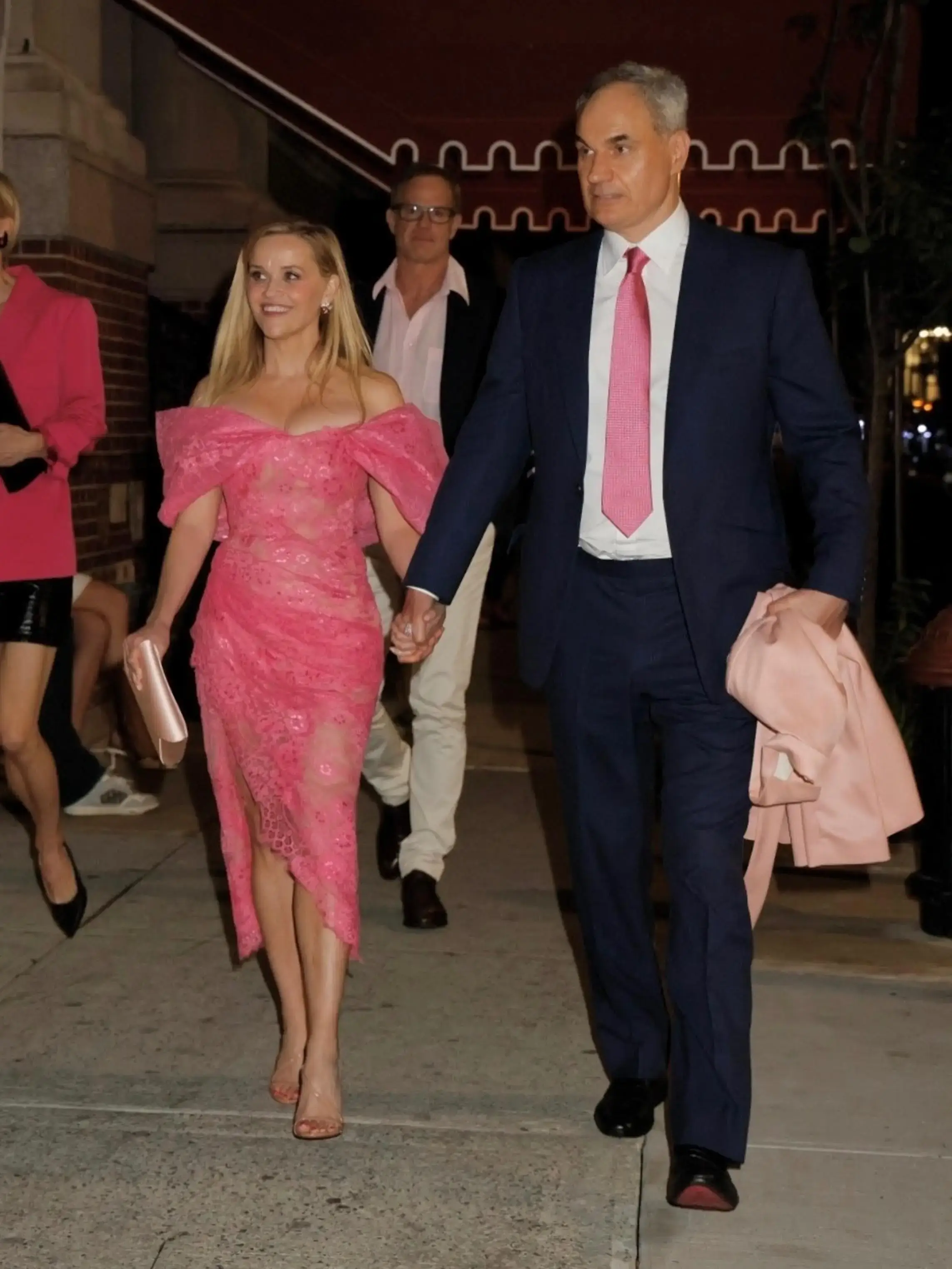 Actress Reese Witherspoon wearing a vibrant off-the-shoulder pink dress walks hand-in-hand down a sidewalk with Oliver Haarmann, who is in a dark blue suit and pink tie.