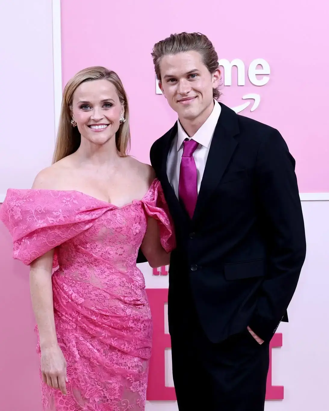 Reese Witherspoon in an off-the-shoulder pink lace dress smiles alongside her son, Deacon Phillippe, who wears a black suit with a magenta tie against a pink background.