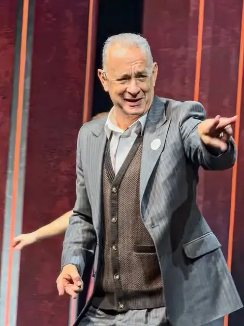 Actor Tom Hanks gestures and points forward while wearing a grey pinstripe suit jacket over a brown cardigan sweater on a stage.