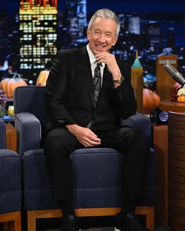 Actor Tim Allen sits smiling in a blue armchair wearing a black suit and a patterned tie on a late-night talk show set.