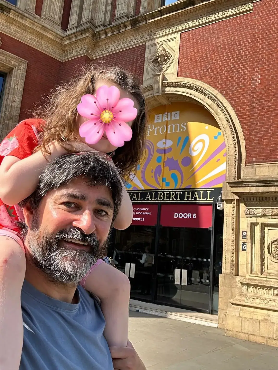 Joplin Sibtain smiles while carrying his young daughter on his shoulders outside the Royal Albert Hall, with her face covered by a pink flower emoji.