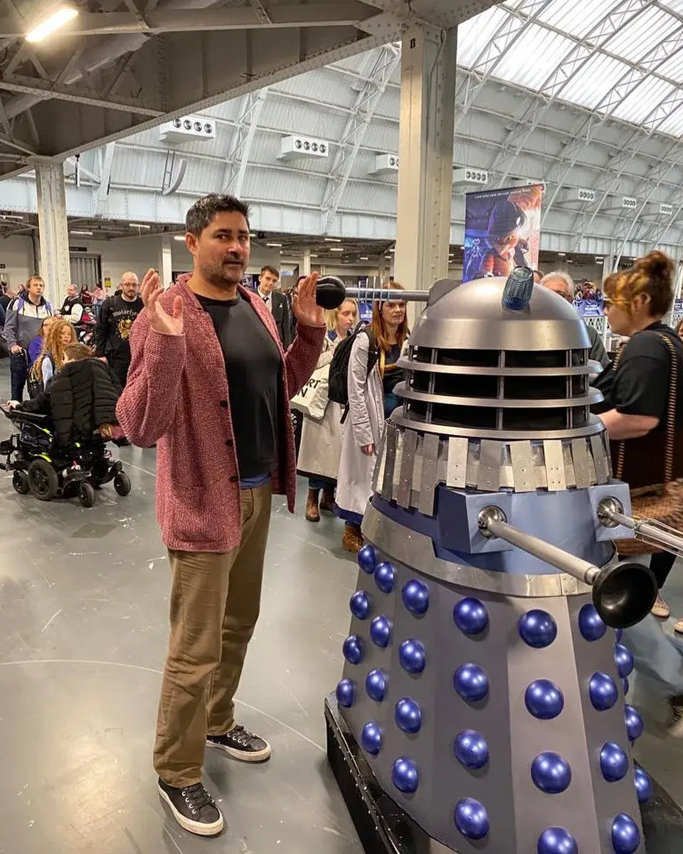 Joplin Sibtain poses playfully with his hands up next to a life-sized Doctor Who Dalek prop at a convention hall.