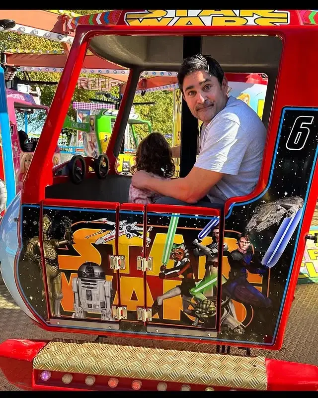 Actor Joplin Sibtain smiles while sitting with his young daughter inside a red Star Wars-themed amusement park ride car.