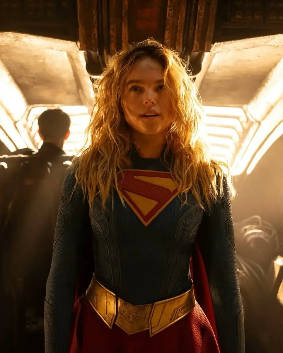 A blonde woman stands in a dimly lit, backlit corridor wearing a Supergirl costume with a prominent "S" insignia.