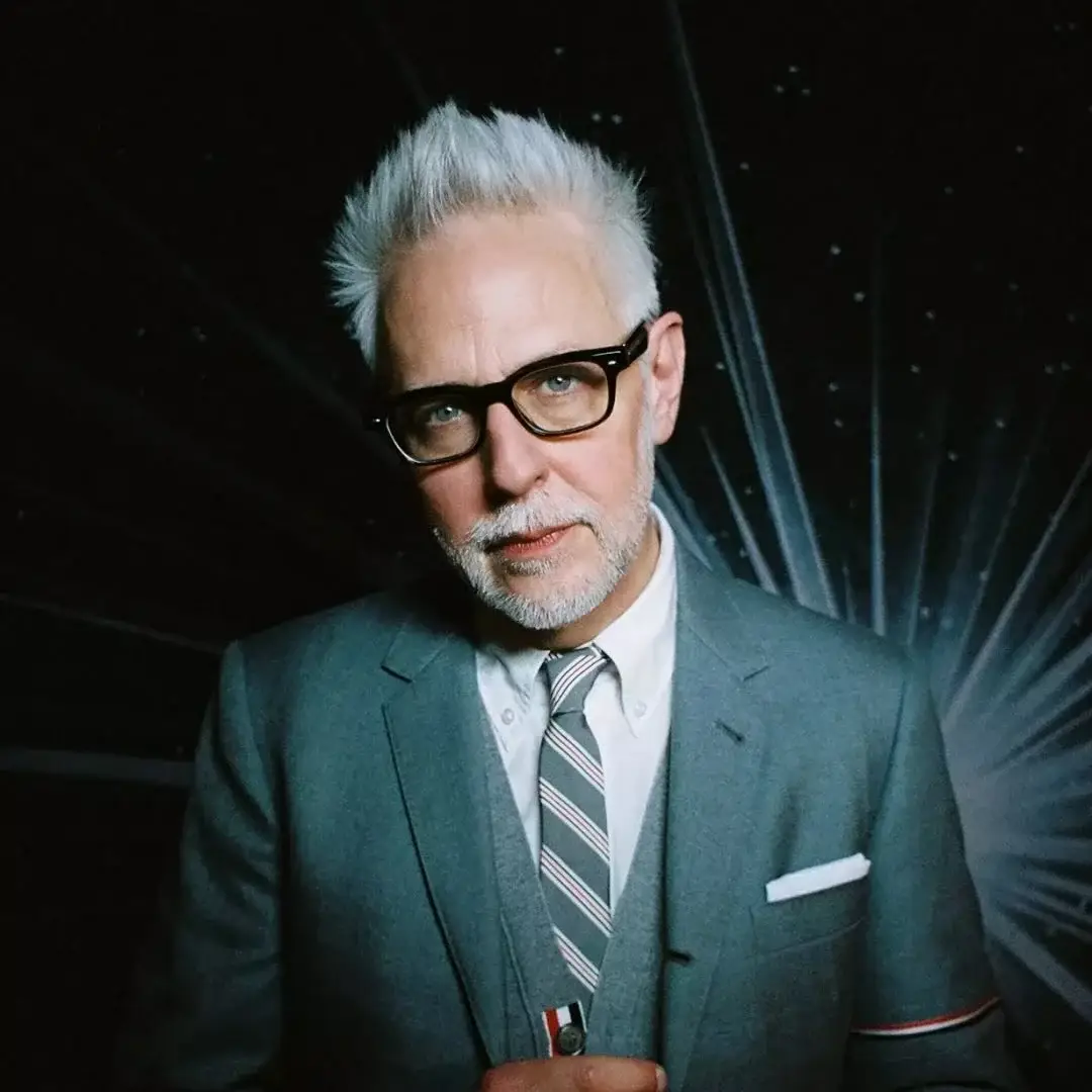 DC Studios Co-CEO James Gunn poses in a grey suit, striped tie, and thick black glasses against a dark background with white light rays.