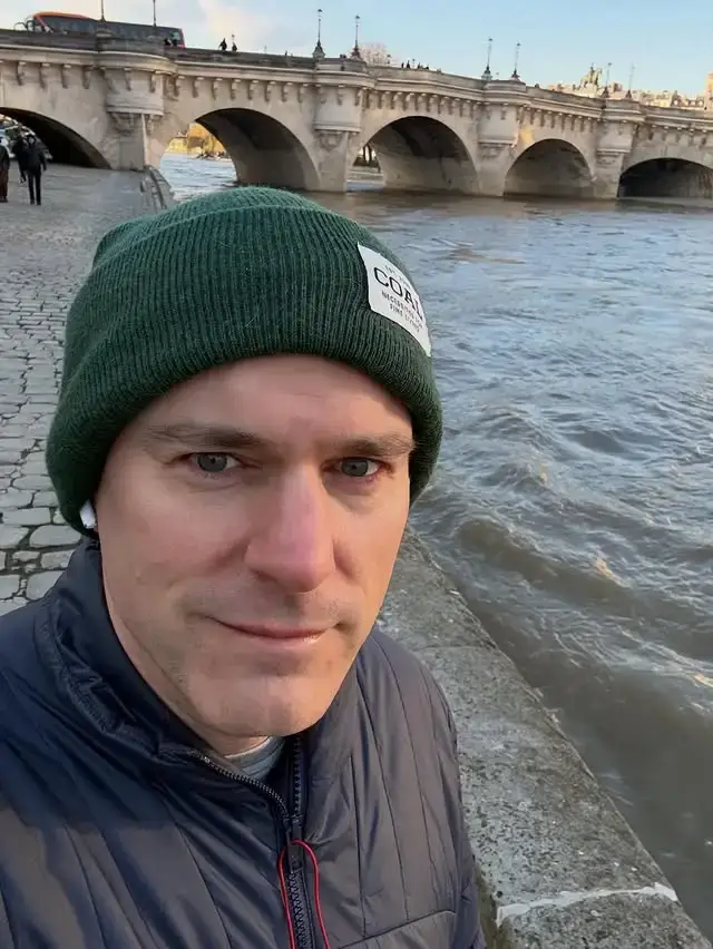 Comic book writer Tom King takes a selfie wearing a green beanie along a riverbank with a stone arch bridge in the background.