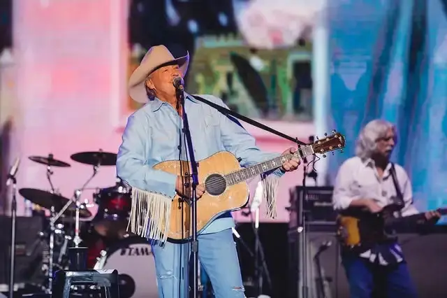 Country singer Alan Jackson sings into a microphone while playing an acoustic guitar on stage with a band behind him.