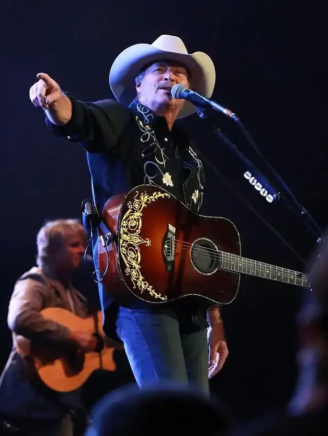 Country star Alan Jackson points to the crowd while holding an ornate acoustic guitar and singing into a microphone on a dimly lit stage.