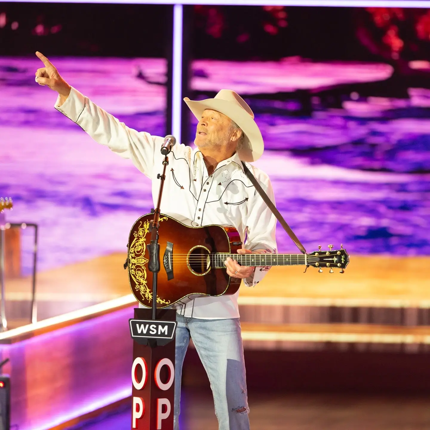 Country artist Alan Jackson wears a white western shirt and cowboy hat while pointing upward on stage behind a WSM Grand Ole Opry microphone stand.